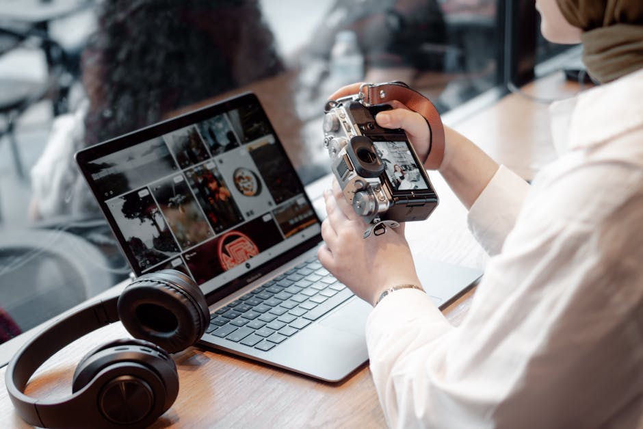 A woman editing photos on her laptop while holding a camera, with headphones nearby on a desk.